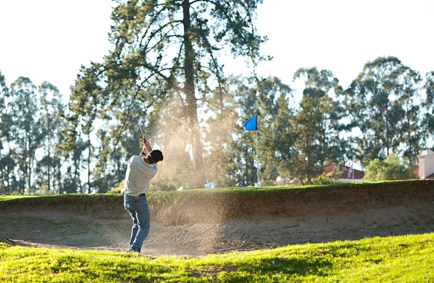 Golfer taking a swing in sand trap, blue flag over golf course hole in background
